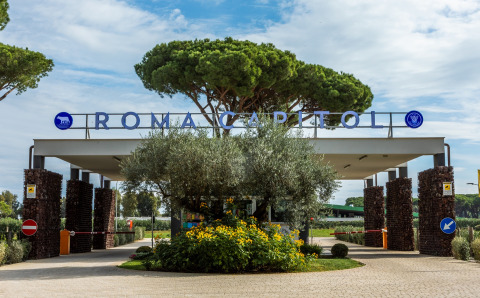 Entrada al Camping Village Roma Capitol en Italia, rodeada de árboles y flores bajo un cielo azul.