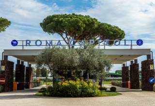 Indgang til Camping Village Roma Capitol i Italien, omgivet af træer og blomster under en blå himmel.