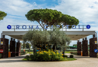 Entrance to Camping Village Roma Capitol in Italy, surrounded by trees and flowers under a blue sky.
