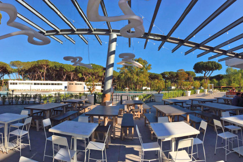 Zona pranzo all'aperto con tavoli e sedie moderne al Camping Village Roma Capitol, circondato da pini.