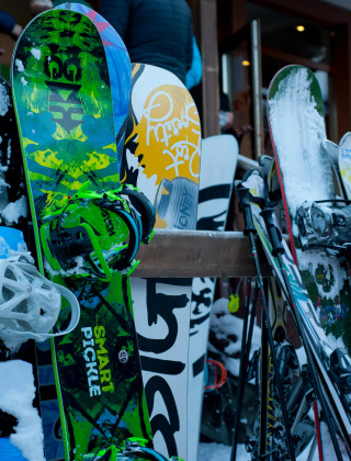 Snowboards und Skier stehen im Schnee draußen an einem Holzständer, nahe einer Treppe und einem Gebäude.