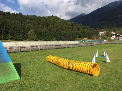 Terrain d'agility canin dans un parc de vacances avec glamping, montagnes et verdure en arrière-plan.