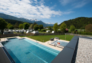 Blick auf einen Campingplatz mit Pool, Wohnwagen, grüner Wiese und Bergen im Hintergrund an einem klaren Tag.