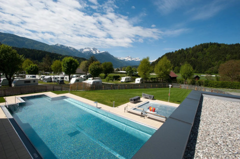 Blick auf einen Campingplatz mit Pool, Wohnwagen, grüner Wiese und Bergen im Hintergrund an einem klaren Tag.