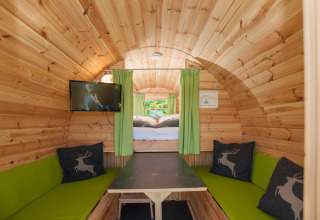 Intérieur d'une cabane de glamping en bois, avec rideaux verts, téléviseur et coussins, dans un parc vacances.