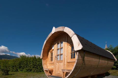 Barrel-shaped wooden glamping pod with windows and door, set in a holiday park on a sunny day.