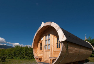 Glamping-tøndeformet hytte i træ under en klar blå himmel på en feriepark omgivet af grønne marker.