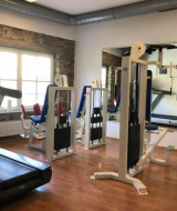 Kleine fitnessruimte in een glampingpark met krachttoestellen, loopband, houten vloer en grote ramen.