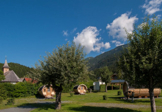 Glamping på feriepark med tøndeformede hytter, bjerge, blå himmel og kirketårn i baggrunden.