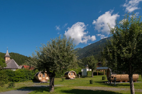 Glamping på feriepark med tøndeformede hytter, bjerge, blå himmel og kirketårn i baggrunden.