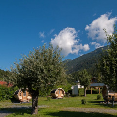 Vakantiepark met glamping in houten tonhutten, bergen, blauwe lucht en kerktoren op de achtergrond.