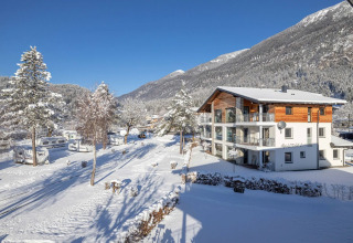 Vista invernal de un parque de glamping con lodge moderno, caravanas y montañas cubiertas de nieve al fondo.