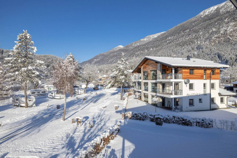 Winter scene of a glamping holiday park with a lodge, caravans, and snowy mountains under a clear blue sky.