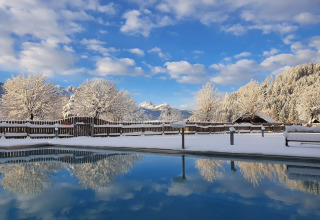 Vue hivernale d’un parc de glamping avec arbres enneigés, montagnes et piscine reflétant le ciel bleu.