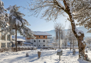 Parque de vacaciones cubierto de nieve con alojamientos glamping, árboles y montañas en un día soleado de invierno.