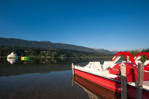 Vista de un lago tranquilo con botes de pedales, montañas y juegos inflables en un parque de glamping.