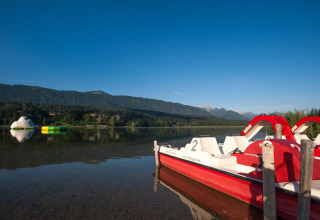 Blick auf einen ruhigen See mit Tretbooten, Bergen und Wasserspielzeug bei einem Glamping-Ferienpark.