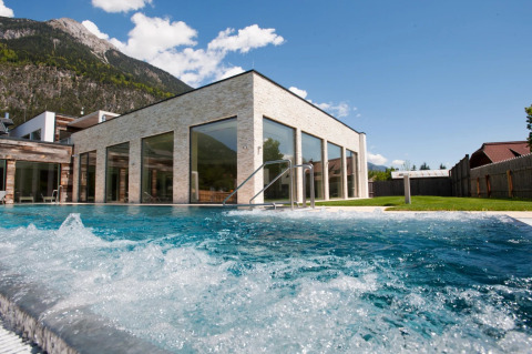 Piscina exterior en un parque de vacaciones moderno con fachada de cristal y montañas de fondo.