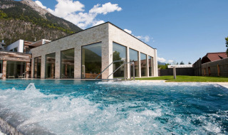 Buitenzwembad bij een modern vakantiepark met glazen gevels en uitzicht op bergen en grasveld.