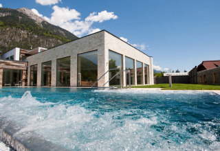 Piscina exterior en un parque de vacaciones moderno con fachada de cristal y montañas de fondo.