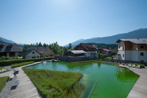 Vue sur une piscine naturelle entourée de terrasses en bois et d’hébergements dans un parc de vacances en montagne.