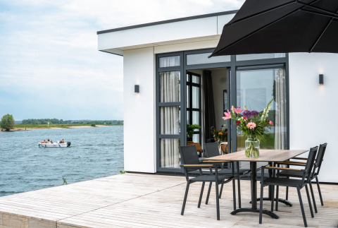 Villa moderna frente al lago con terraza, mesa de comedor, jarrón de flores y bote en el agua de fondo.