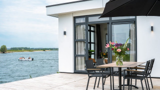 Villa moderna frente al lago con terraza, mesa de comedor, jarrón de flores y bote en el agua de fondo.