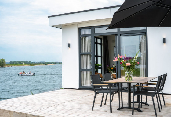 Villa moderna frente al lago con terraza, mesa de comedor, jarrón de flores y bote en el agua de fondo.