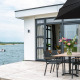 Moderne villa ved vandet med terrasse og spisebord, blomster i vase, udsigt over sø og båd i baggrunden.