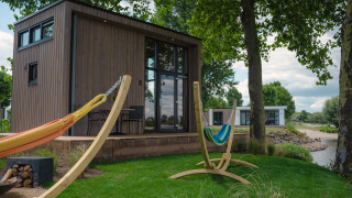 Pequeña casa de madera junto al río con hamacas y terraza en MarinaPark Bad Nederrijn, rodeada de árboles.