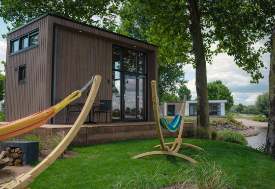 Pequeña casa de madera junto al río con hamacas y terraza en MarinaPark Bad Nederrijn, rodeada de árboles.