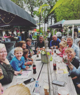 En gruppe mennesker hygger sig ved et langt bord på Camping Emmen i Drenthe, Holland, omgivet af natur.