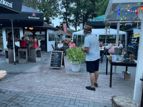 Outdoor café at Camping Emmen in Drenthe, Netherlands, with guests, a menu board, and festive lights.