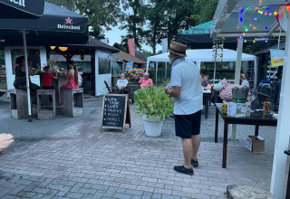 Outdoor café at Camping Emmen in Drenthe, Netherlands, with guests, a menu board, and festive lights.