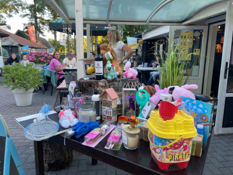 Outdoor scene at Camping Emmen in Drenthe, Netherlands, with guests and a table displaying various raffle prizes.