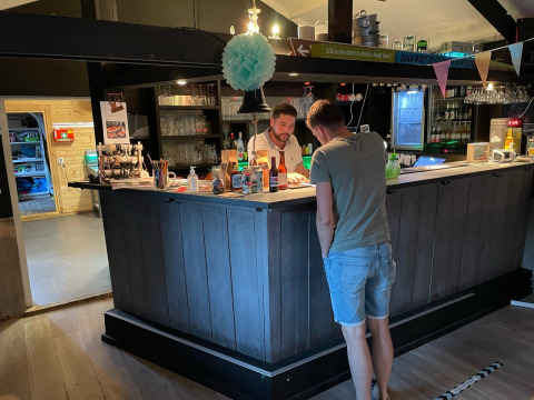 Twee mannen praten aan een gezellige bar in een lodge, met houten interieur, flessen en glazen op de plank.