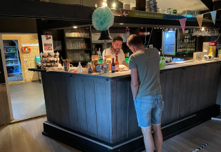 Deux hommes discutent à un bar confortable de lodge avec déco bois, bouteilles et verres en arrière-plan.