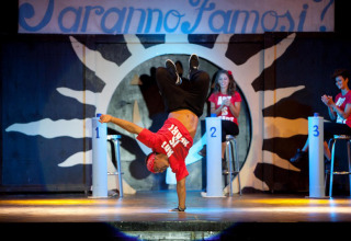 Een breakdancer voert een act uit op een podium in een vakantiepark op Sardinië, Italië, terwijl juryleden kijken.