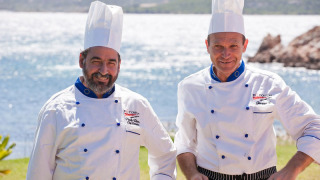 Dos chefs con uniforme blanco y gorros sonríen junto al mar en Centro Vacanze Isuledda, Cerdeña.