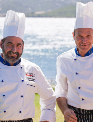 Dos chefs con uniforme blanco y gorros sonríen junto al mar en Centro Vacanze Isuledda, Cerdeña.
