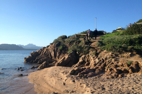 Strandzicht aan Centro Vacanze Isuledda, Sardinië met rotsen, zee en mensen wandelend op het pad.