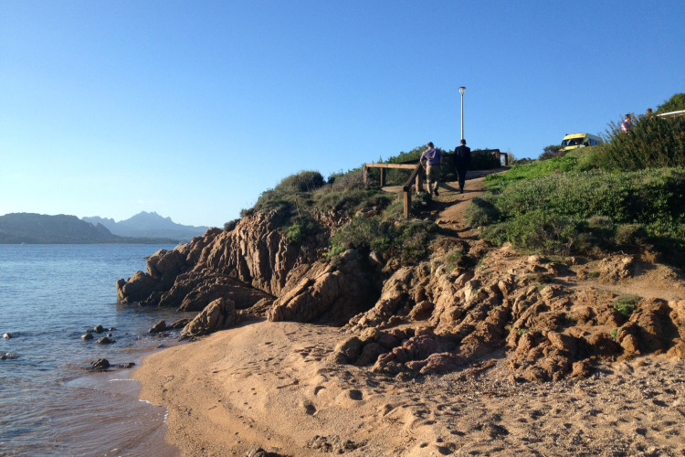Beach view at Centro Vacanze Isuledda, Sardinia showing rocks, sea, sandy shore and people on a footpath.