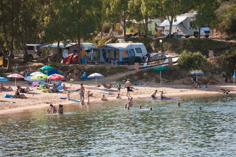 Feriecampingplads i Sardinien med familier, campingvogne og telte ved stranden samt folk der bader.