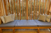 Banco de madera al aire libre con cojines grises y almohadas estampadas en SunLodge Sabai, rodeado de plantas.