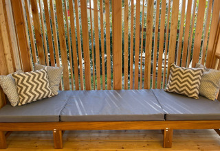 Banco de madera al aire libre con cojines grises y almohadas estampadas en SunLodge Sabai, rodeado de plantas.