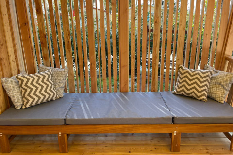 Banco de madera al aire libre con cojines grises y almohadas estampadas en SunLodge Sabai, rodeado de plantas.