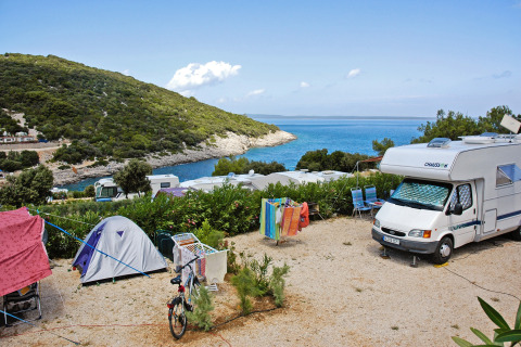 Camping en la costa de Croacia con tiendas, autocaravanas y vistas al mar y colinas verdes al fondo.