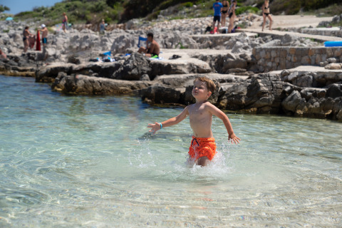 Bambino in costume arancione gioca nell’acqua limpida al Camping Village Poljana, Croazia, giornata di sole.
