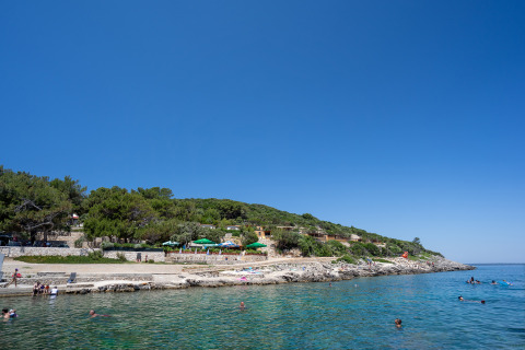 Kustzicht aan Camping Village Poljana, Kroatië, met zwemmers, groene heuvels en heldere blauwe hemel.