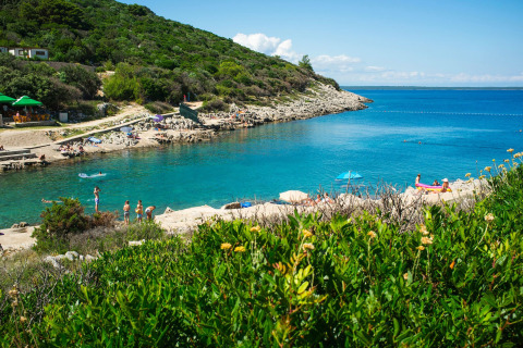 Camping Village Poljana en Croacia con visitantes disfrutando de agua azul clara, colinas y playa rocosa.